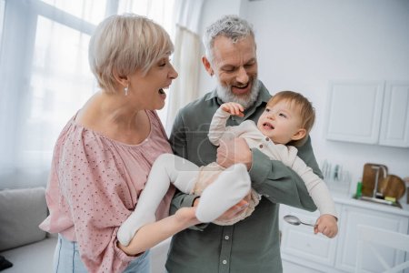 Téléchargez les photos : Joyeux couple d'âge moyen s'amuser avec la petite-fille souriante tenant cuillère dans la cuisine - en image libre de droit