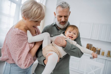 Photo for Carefree baby girl holding spoon near smiling grandparents in kitchen - Royalty Free Image
