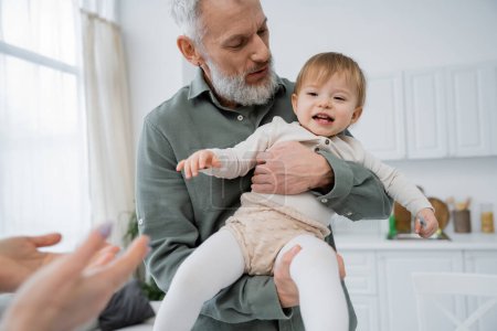 mature bearded man holding cheerful toddler granddaughter in kitchen