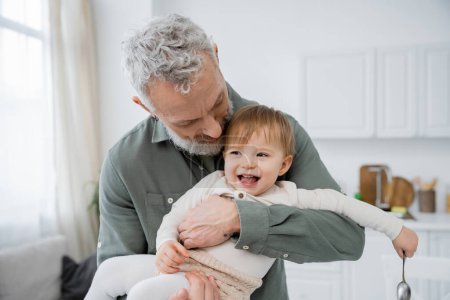 bearded mature man embracing carefree granddaughter holding spoon in kitchen