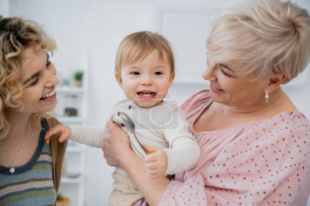 carefree baby girl holding spoon near cheerful mom and granny in kitchen