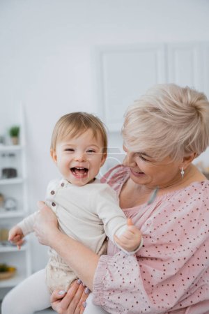 Téléchargez les photos : Réjoui bébé fille tenant cuillère près de grand-mère souriante dans la cuisine - en image libre de droit