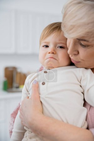 mature woman hugging and calming displeased granddaughter grimacing and looking at camera in kitchen