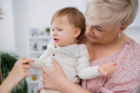 smiling mature woman hugging displeased baby girl in kitchen