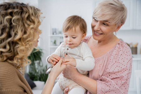 smiling mature woman looking at adult daughter giving spoon to little girl in kitchen
