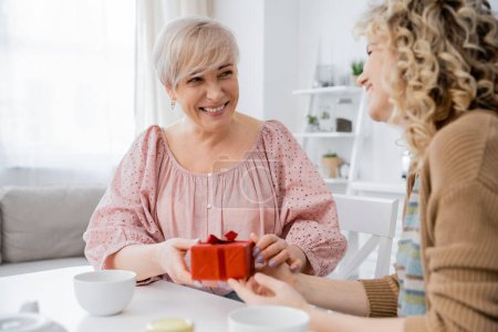 pleased middle aged woman holding present and looking at adult daughter while sitting near tea cups in kitchen