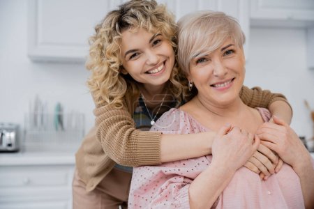 überglückliche Frau mit lockigem blondem Haar umarmt glückliche Mutter und lächelt in die Kamera in der Küche