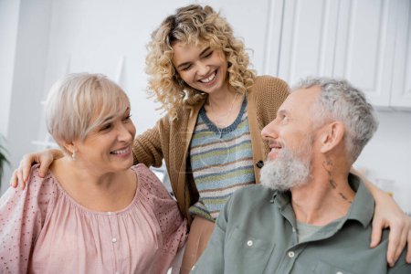 Téléchargez les photos : Femme souriante aux cheveux blonds ondulés embrassant joyeux parents d'âge moyen dans la cuisine à la maison - en image libre de droit
