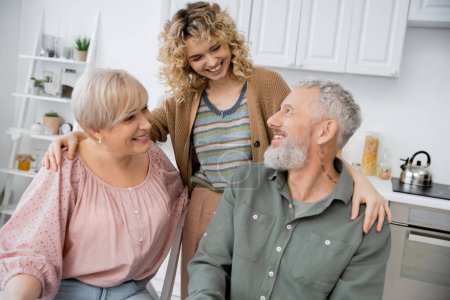 Cheerful woman with curly blonde hair hugging happy parents in kitchen