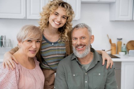 pleased middle aged couple smiling at camera near adult daughter in kitchen at home
