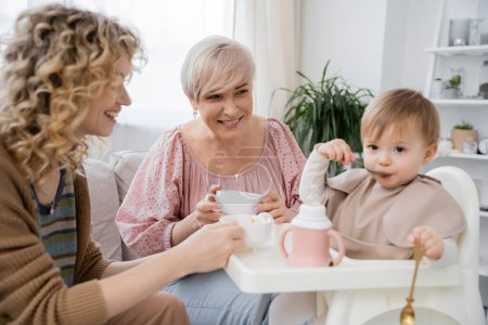 cheerful women looking at baby girl with spoon having breakfast in kitchen
