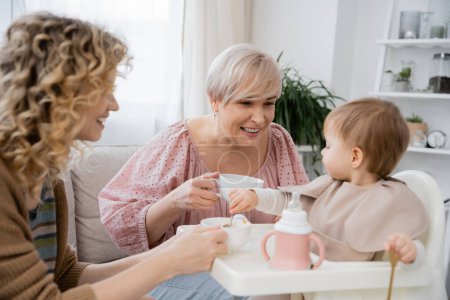 cheerful mature woman holding tea cup and looking at granddaughter having breakfast in kitchen