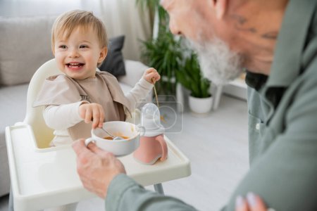 Téléchargez les photos : Fille insouciante avec cuillères assis sur chaise bébé près du bol avec petit déjeuner et grand-père sur le premier plan flou dans la cuisine - en image libre de droit