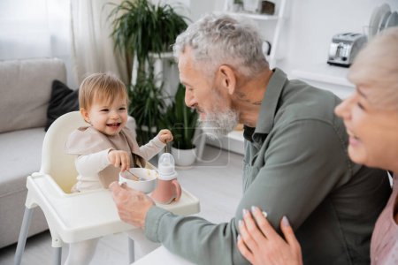 Älteres Paar lächelt in der Nähe unbeschwerte Enkelin mit Löffeln sitzen auf Babystuhl in der Nähe Schüssel mit Frühstück in Küche