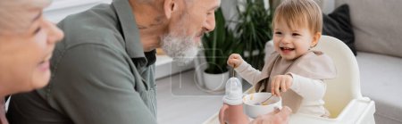 Téléchargez les photos : Fille ravie tenant cuillères tout en prenant le petit déjeuner près de grands-parents heureux dans la cuisine, bannière - en image libre de droit