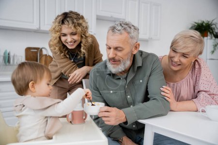 bärtiger Mann sitzt mit Schale neben fröhlicher Familie und Kleinkind-Enkelin mit Löffel in Küche