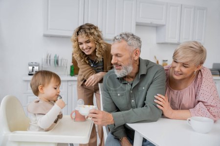 smiling bearded man holding bowl near granddaughter sitting on baby chair during breakfast in kitchen