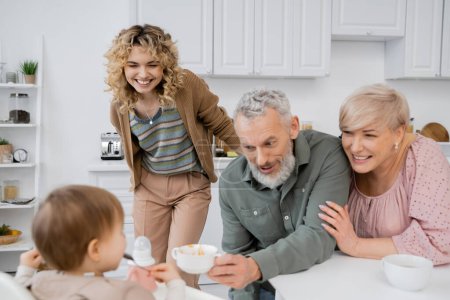 cheerful bearded man holding bowl near blurred granddaughter and smiling family in kitchen