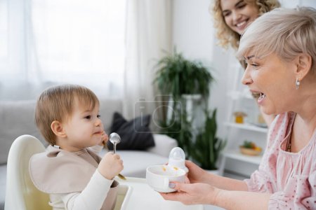 mature woman opening mouth while holding bowl near granddaughter with spoon having breakfast in kitchen