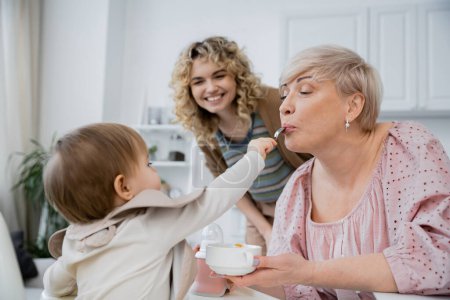 little girl with spoon feeding grandma near smiling mother in kitchen