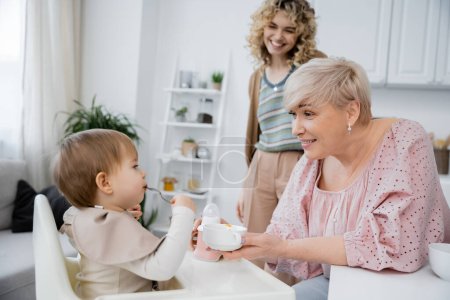 pleased middle aged woman holding bowl near toddler granddaughter with spoon during breakfast in kitchen