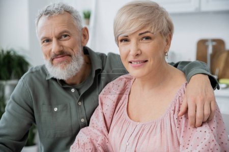 Téléchargez les photos : Heureuse femme d'âge moyen et homme barbu souriant à la caméra tout en étant assis dans la cuisine à la maison - en image libre de droit
