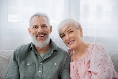 happy bearded man with smiling wife looking at camera at home in living room