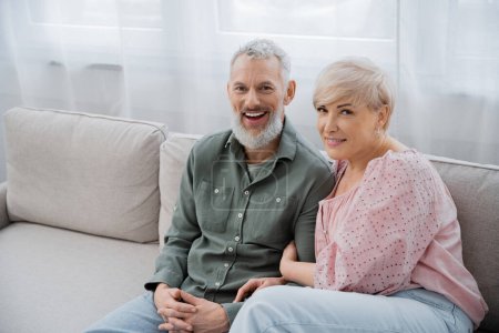 Foto de Mujer de mediana edad con el marido barbudo emocionado sentado en el sofá en la sala de estar y sonriendo a la cámara - Imagen libre de derechos