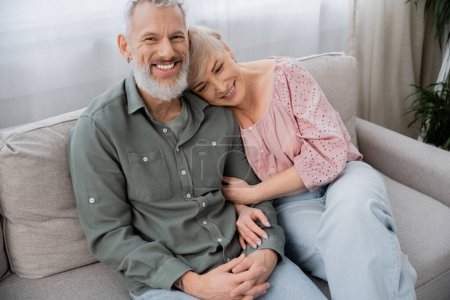 glückliche Frau mittleren Alters stützt sich auf fröhlichen bärtigen Mann lächelnd in die Kamera auf Couch im Wohnzimmer