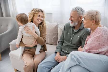 fröhliche blonde Frau hält ihre kleine Tochter in der Hand und schaut glückliche Eltern auf der Couch im Wohnzimmer an