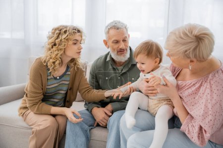 curly woman calming upset baby near mature parents on sofa in living room