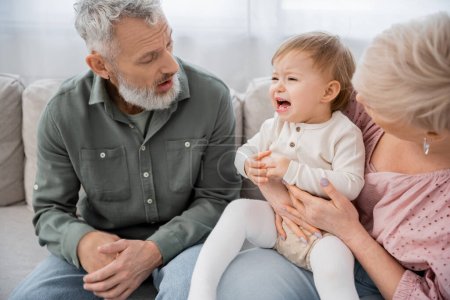 confused middle aged couple calming crying granddaughter on couch in living room
