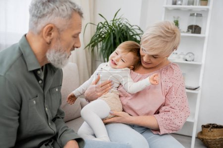 mature woman holding displeased granddaughter while sitting near bearded husband in living room