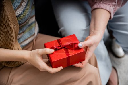 partial view of woman holding red gift box near mature mother while sitting at home