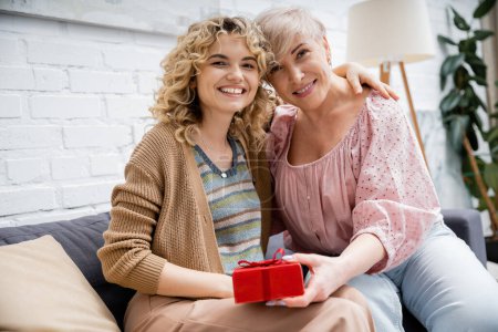 blonde woman with wavy hair embracing happy mother holding gift box while sitting on sofa at home