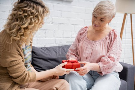 curly woman presenting gift to pleased mother while sitting on couch at home