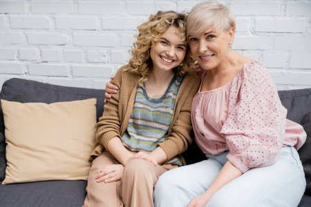 Photo for Happy mature woman with cheerful blonde daughter sitting on couch in living room and looking at camera - Royalty Free Image
