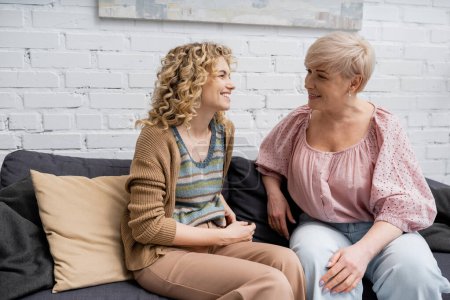 mujer alegre con cabello rubio ondulado hablando con la sonriente madre de mediana edad en el sofá en la sala de estar