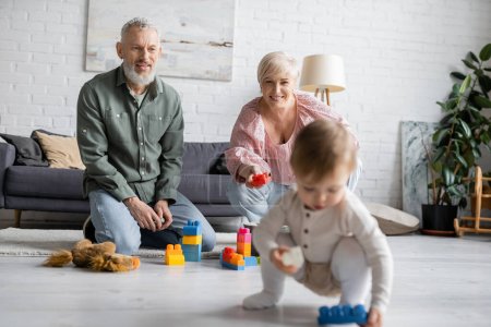 glückliche Großeltern mittleren Alters sitzen auf dem Boden in der Nähe von Bauklötzen, während kleines Mädchen mit Spielzeugauto im Wohnzimmer spielt