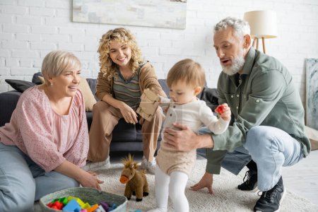Lächelnde Frauen schauen bärtige Männer an, die mit Enkelin auf dem Fußboden im Wohnzimmer spielen