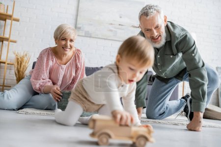 überglückliches Paar mittleren Alters blickt auf verschwommene Enkelin, die mit Spielzeugauto auf dem Boden im Wohnzimmer spielt