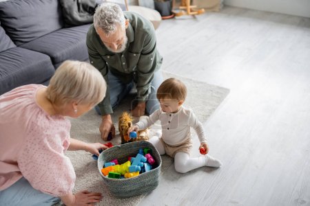 high angle view of mature couple and toddler girl near wicker basket with toys on floor in living room