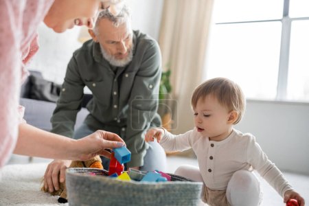 Kleines Mädchen schaut sich Spielzeug im Weidenkorb an, während es mit den Großeltern im Wohnzimmer spielt