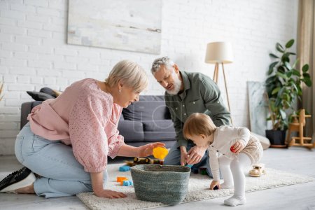Paar mittleren Alters spielt mit Kleinkind-Enkelin auf Bodenteppich im modernen Wohnzimmer