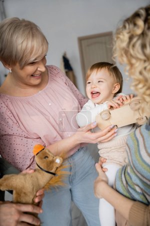 Sorgloses Kind hält Spielzeugauto in der Hand und lacht neben Großeltern und Mutter im Wohnzimmer