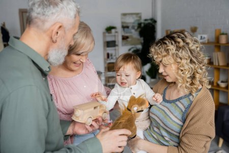 middle aged grandparents holding toys near toddler granddaughter at home