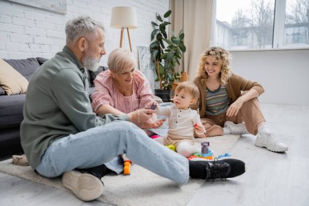 fröhliches reifes Paar spielt Bauklötzchenspiel mit Enkelin, während es im modernen Wohnzimmer auf dem Boden sitzt