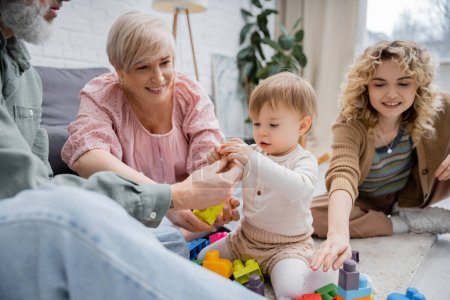 smiling grandparents giving toys to toddler girl while playing together in living room