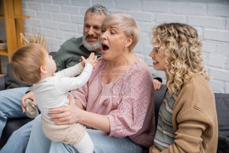 Téléchargez les photos : Grand-mère ouverture bouche tout en s'amusant avec tout-petit fille sur canapé dans le salon - en image libre de droit
