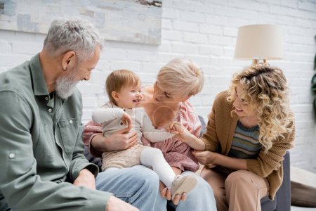 Foto de Niña emocionada riendo cerca de la madre sonriente y abuelos sentados en el sofá en la sala de estar - Imagen libre de derechos